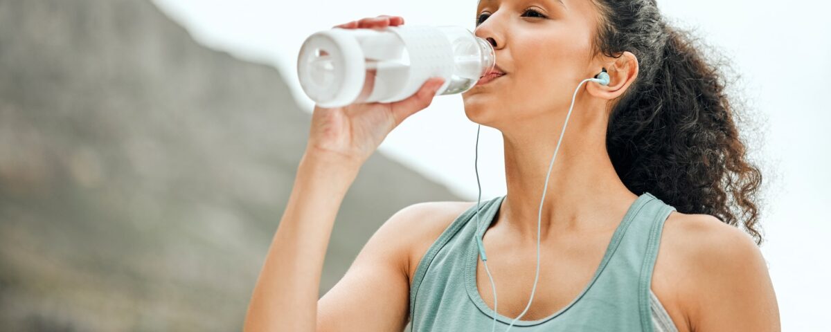 Young woman with a long dark ponytail drinks from a clear water bottle outdoors while wearing a seafoam green tank top and corded headphones. Photo is take from the torso up and the mountains are blurred in the background. The woman gives an impression of post workout.