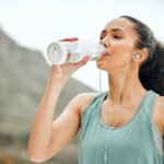 Young woman with a long dark ponytail drinks from a clear water bottle outdoors while wearing a seafoam green tank top and corded headphones. Photo is take from the torso up and the mountains are blurred in the background. The woman gives an impression of post workout.